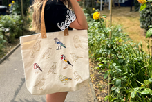 A person outdoors carrying a large cream canvas tote bag with embroidered bird illustrations and tan shoulder straps. The background shows a garden path and greenery.