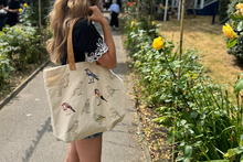 A woman in a garden wearing a black t-shirt and carrying a cream canvas tote bag with various bird illustrations. Some of them have been embroidered in colour. The background shows green foliage and yellow flowers.